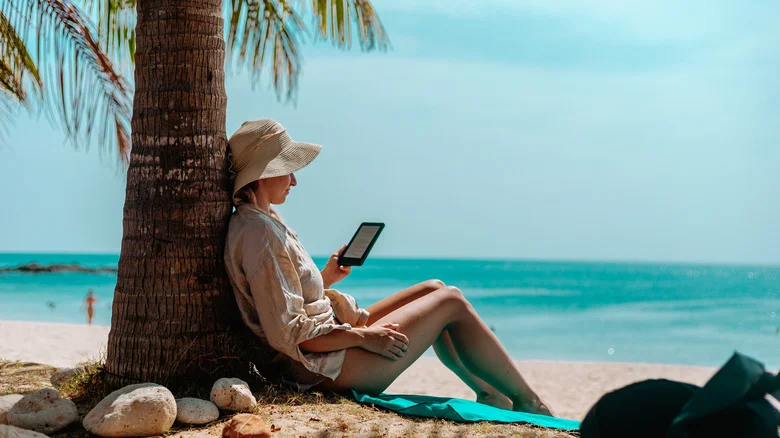 A woman sits on a beach against a palm tree reading from her Kindle.
