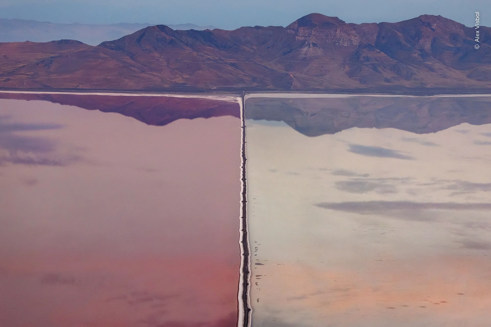 A single engineered line reshaping an entire wetland, waters perfectly still reflecting the mountainous backdrop
