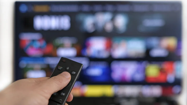 Hand holding a remote in front of a blurred smart TV in the background.