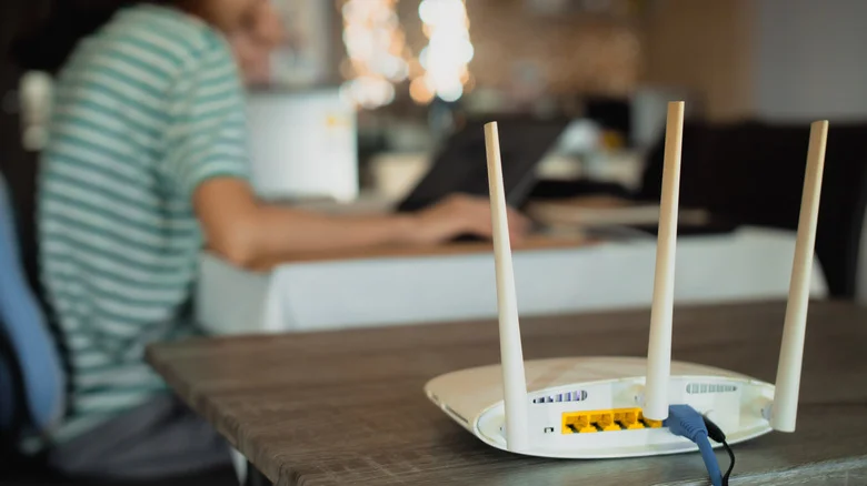 A Wi-Fi router with an Ethernet cable plugged in, with a person working on a laptop blurred in the background.